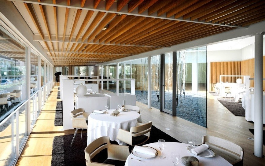 Interior del restaurante con techo de lamas, alfombras grises y patio acristalado con árboles